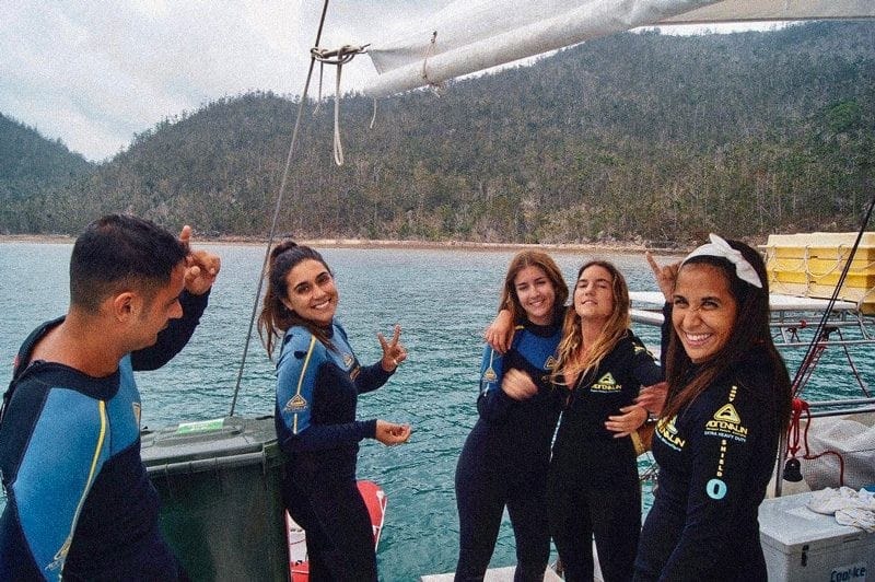 grupo de chicas en traje de submarinismo encima de un barco sonriendo a cámara y detrás el paisaje de las islas Whitsundays